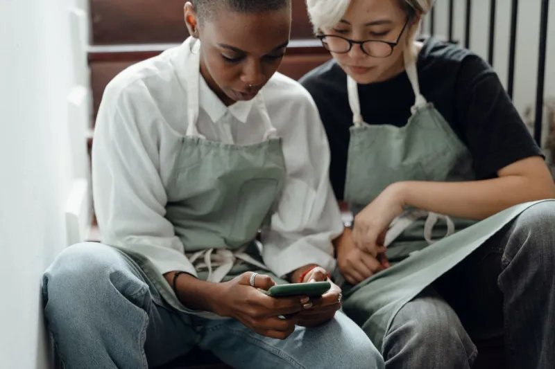 two ladies sat on a staircase wearing aprons looking at a mobile phone screen